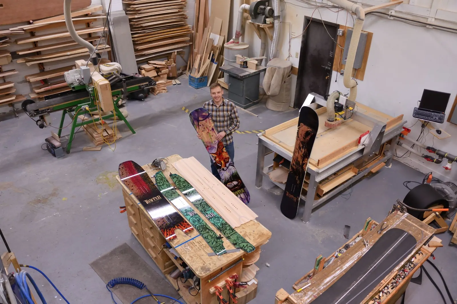 Greg in the Trapper Snowboards shop, holding a finished board beside the CNC router, with boards in progress on the bench